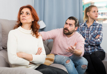 Sad mature female sitting after quarrel and daughter with boyfriend on background at home