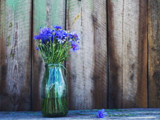 Bright blue bouquet of cornflowers in an old glass bottle on the background of an old wooden board, close-up, copy space. Summer vintage bouquet of meadow herbs