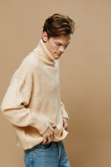 a handsome, slender guy in a long oversized beige sweater straightens his clothes while standing on a plain background