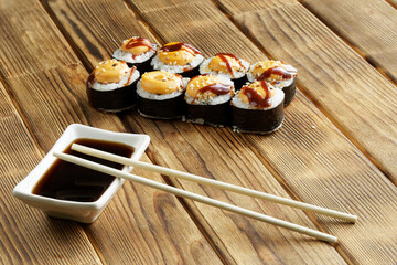 Appetizing rolls with rice, nori, fish and spices next to the Japanese gravy boat - seyuzaru with chopsticks - varibashi and soy sauce. Table made of natural pine boards. Selective focus.