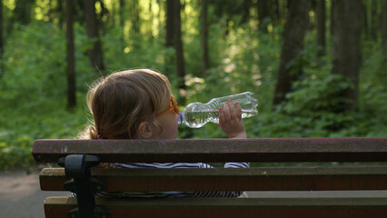 View from back cheerful little girl in striped t-shirt and orange glasses drinks clean ecological...