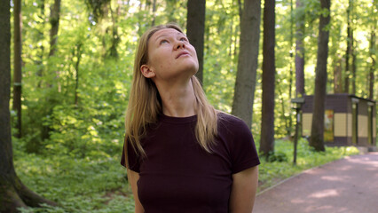 Fototapeta premium beautiful slender blonde walks through picturesque green forest at sunset or dawn. woman in black t-shirt looks up at birds or treetops. lady on walk in forest walks alone along paths.