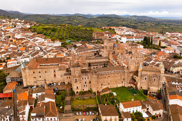 Fototapeta na wymiar Gothic-Mudejar style building of Royal Monastery of Saint Mary in Spanish town of Guadalupe, located in green valley of province of Caceres overlooking brownish roofs of houses, as seen from drone..