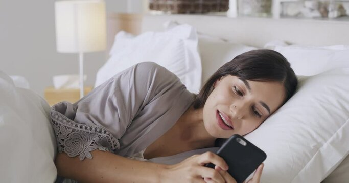 A young woman using her phone while lying in her bed. Curious female reading interesting post online, reacting or reply to a message on social media. Excited woman checking notifications for good news