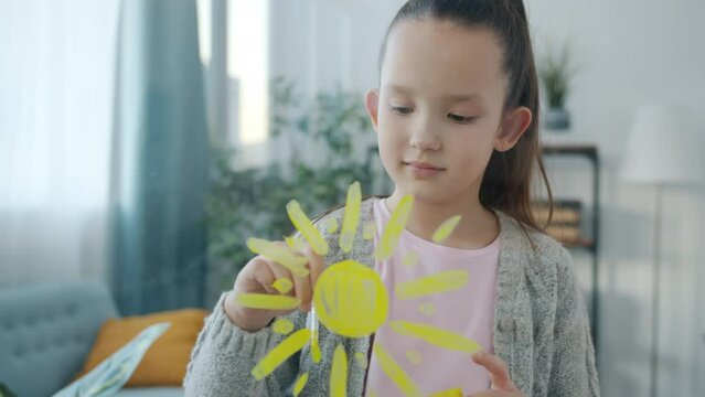 Portrait of lovely kid painting sun with painbrush on glass surface in apartment while girl is enjoying art in leisure time alone. Childhood and hobby concept.