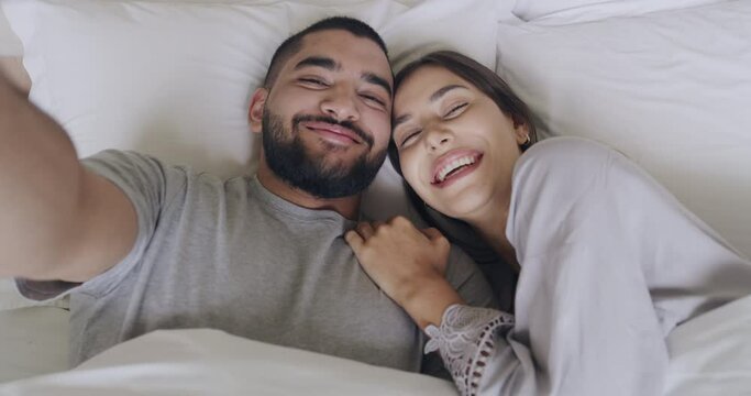 Goofy young interracial couple making funny faces and being silly while taking a selfie in bed. Playful lovers laughing and having fun while laying together. Sticking out tongue and looking cross eyed