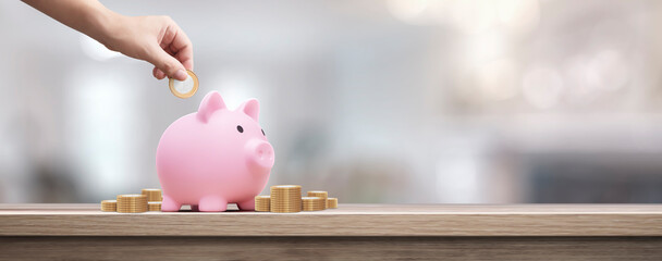 man depositing coins in a pink piggy bank on a wooden table - savings concept - obrazy, fototapety, plakaty