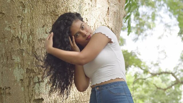 Sexy young latina model playing in her curly hair at the base of a tree trunk