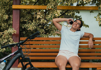 latina girl, sitting on a park bench with her bicycle on one side, answers a call on her phone, with a big expression and a smile on her face. 