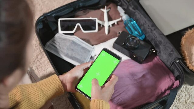 Woman using smartphone blank green screen and packing suitcase bag for travel trip weekend vacation at home, female check information in tourism website on mobile phone before go to airport