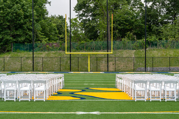 Fototapeta premium White chairs set-up in rows on a green synthetic turf athletic field for a high school graduation ceremony. 
