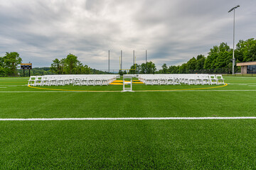 White folding chairs set-up in rows, with single chair at center, on a green synthetic turf athletic field for a high school graduation ceremony. 