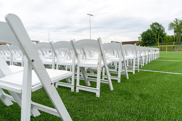 White chairs set-up in rows on a green synthetic turf athletic field for a high school graduation ceremony. 
