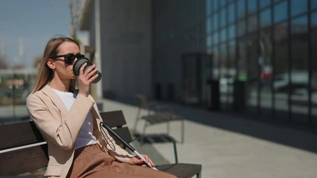 Visually impaired woman sitting on bench near business center and drinking hot coffee. Caucasian young female in black eyeglasses and casual wear relaxing outdoors.