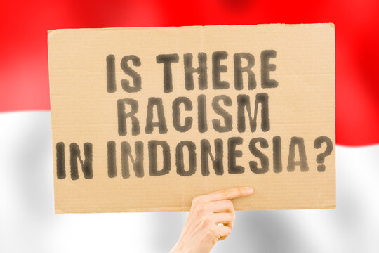 The question " Is there racism in Indonesia? " is on a banner in men's hands with a blurred Indonesian flag in the background. Interracial. Issues. Justice. Nation. Politics. Peace. Patriotism
