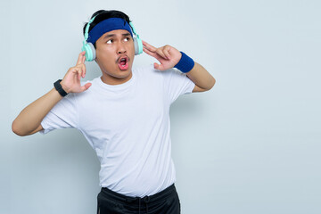 Shocked young asian man sporty fitness trainer instructor in blue headband and white t-shirt.   Listen to music with headphones isolated over white background