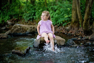 Cute little toddler girl having fun by a river on warm and sunny summer day. Happy excited preschool child splashing with water in forest stream creek.