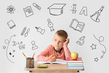 Thoughtful little schoolboy sitting at desk against light background
