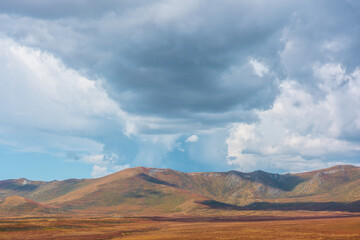 Motley autumn landscape with sunlit high mountain plateau and mountain range under dramatic sky. Vivid autumn colors in mountains. Sunlight and shadows of clouds in changeable weather. Sunny and rainy