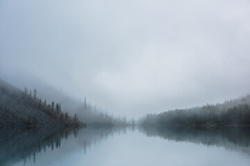 Tranquil meditative misty scenery of glacial lake with pointy fir tops reflection at early morning. Graphic EQ of spruce silhouettes on calm alpine lake horizon in mystery fog. Ghostly mountain lake.