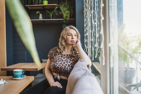Attractive chubby young blonde woman in a leopard blouse drinks coffee from a turquoise cup in a cafe sits near a panoramic window. Plus size girl with a cup have fun 