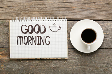 Notebook with text GOOD MORNING and cup of coffee on wooden background
