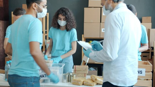 Group of charity workers in face masks and gloves packing groceries for donation during pandemic. Occupation and humanitarian aid concept.