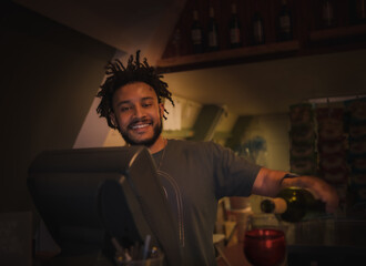 Portrait of handsome African American bartender at work pouring red wine; dim light
