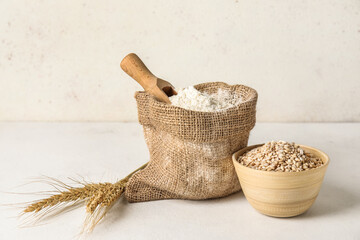 Bag with flour and bowl with wheat on light background