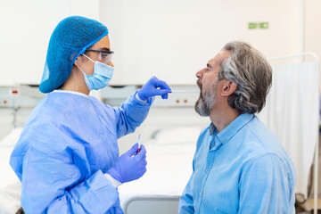 A mature Caucasian man in a clinical setting being swabbed by a healthcare worker in protective...