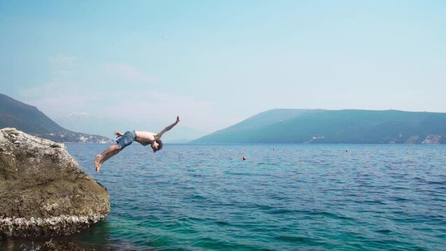 Caucasian young boy jumping into sea from a rock in slow motion doing somersault