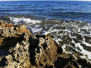Long-hardened lava, porous, sharp coast of the Mediterranean Sea with oncoming waves against a sky with clouds.