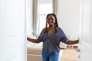 Fototapeta na wymiar Welcome. Portrait of cheerful African woman inviting visitor to enter his home, happy young woman standing in doorway of modern apartment showing living room with hand