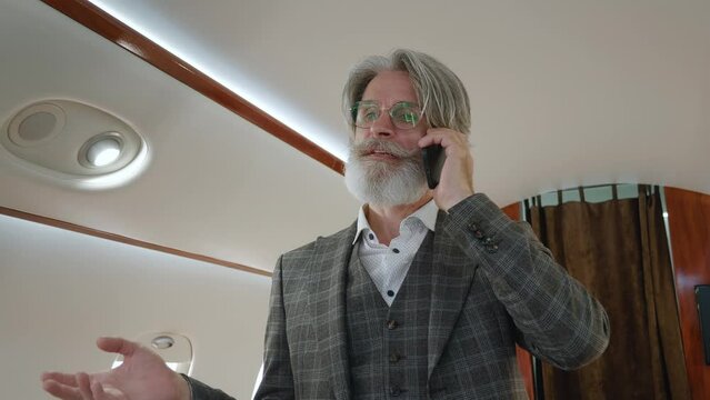 Portrait of stylish senior millionaire man talking on mobile phone, standing in private airplane. Smiling bearded CEO businessman having phone call in luxury private jet.