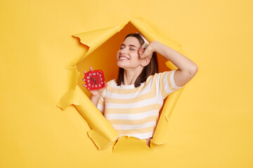 Portrait of satisfied young Caucasian woman wearing striped shirt and hair ban d posing in yellow paper hole, standing and stretching body after sleeping, wakes up in good mood, holding alarm clock.