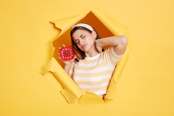 Portrait of sad upset sleepy woman wearing striped shirt and hair band posing in yellow paper hole,...