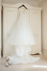 Fototapeta na wymiar A white beautiful bride's dress hangs on the door of a wooden wardrobe in a bright white room.