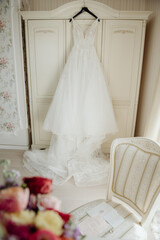 A white beautiful bride's dress hangs on the door of a wooden wardrobe in a bright white room.