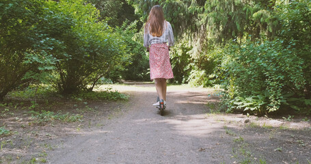 Female in dress rides on an electric scooter along a path in a park with green trees. Eco concept, green energy, vehicle. Rear view, daytime, spring summer.