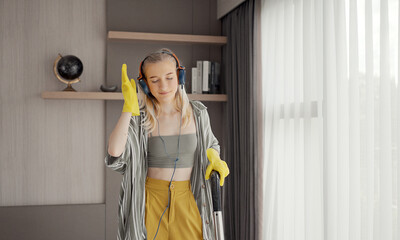 Young girl havig fun while cleaning floor with vacuum cleaner. Happy woman doing housework at home enjoy music wearing earphones.