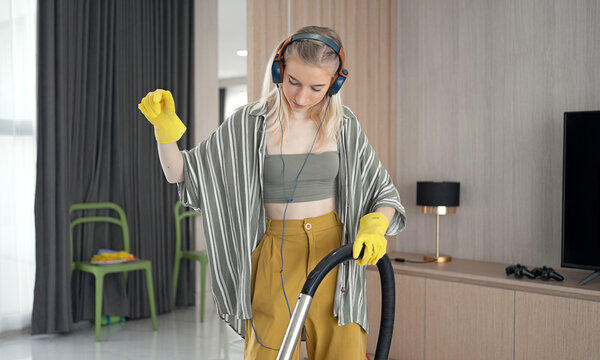 Young girl havig fun while cleaning floor with vacuum cleaner. Happy woman doing housework at home enjoy music wearing earphones.