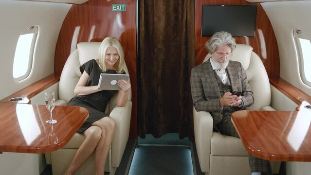 Mature rich couple using smartphone and tablet pc while luxury traveling in private jet plane. Senior man and woman using gadgets, looking at window while flying on first class airlines