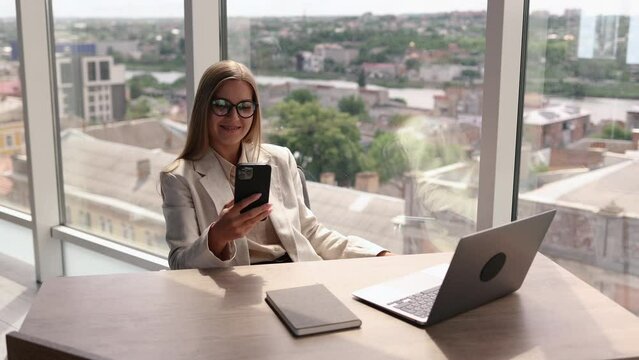 Smiling young business woman professional talking on phone using laptop sit at home office desk, happy female customer make mobile call confirming online website shopping order delivery concept