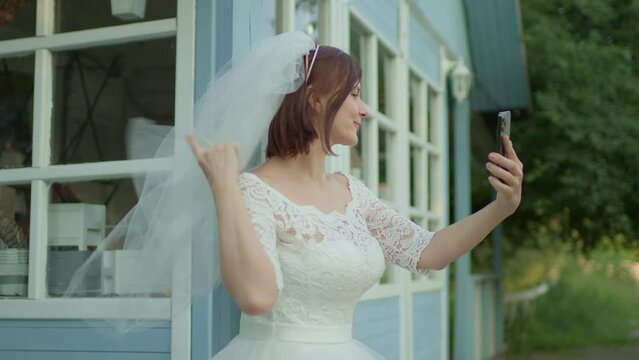 30s bride in white wedding dress taking selfie photos standing near rural house. Happy newlywed enjoys mobile picturing. Slow motion, handheld camera.