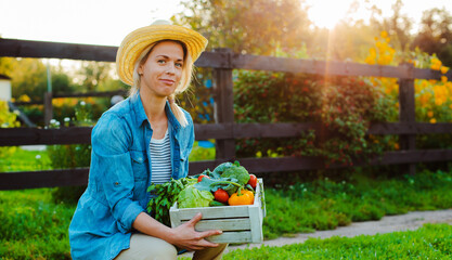 Young 30-35 years old beautiful Woman farmer in hat with box of fresh ecological vegetables on garden background at sunset.