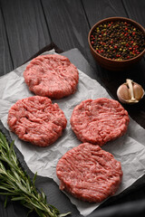 Raw beef burger patties on butcher's wooden board, rosemary and pepper. Black background. View from above.