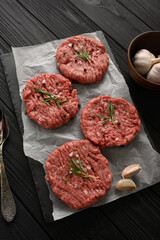 Raw beef burger patties on butcher's wooden board, rosemary and pepper. Black background. View from above.
