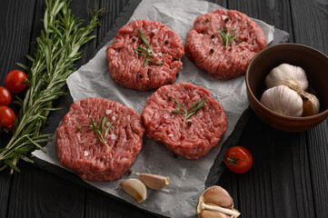 Raw beef burger patties on butcher's wooden board, rosemary and pepper. Black background. View from above.