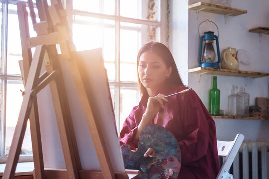 Painter paints with watercolors in workshop at home. Portrait of pretty young woman artist in art studio and painting on canvas