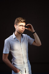 Obraz premium Handsome young man in early 20s wearing smart casual clothes and adjusting eyeglasses while looking away, standing against black background.